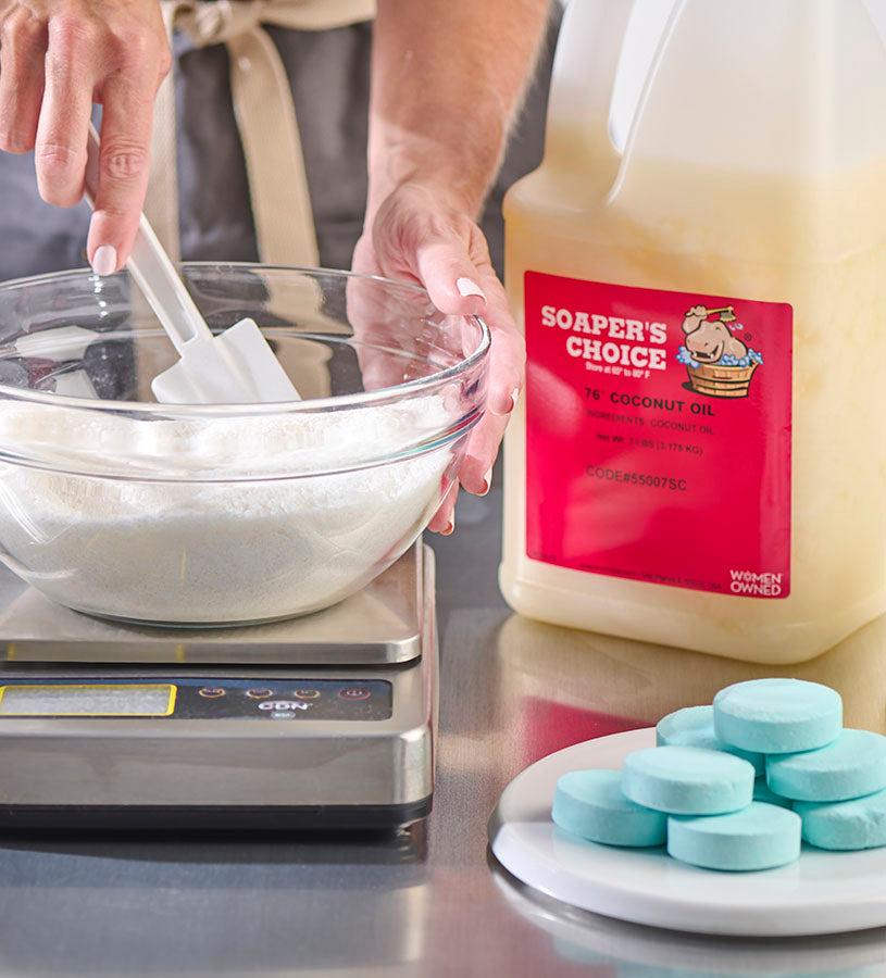 Hands mixing white soap base in a glass bowl on a scale, with Soaper's Choice coconut oil container and finished turquoise soap bars on a plate nearby